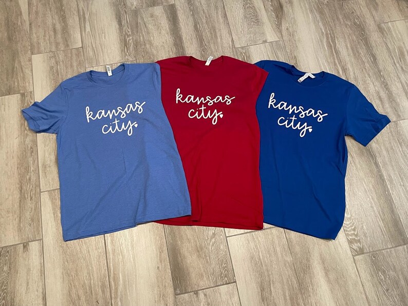 May include: Three t-shirts with the text "kansas city" in white script on a blue, red, and royal blue background.
