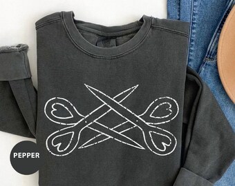 May include: A dark gray sweatshirt with a white graphic of a pair of scissors with heart-shaped handles. The word "PEPPER" is printed in white on the bottom left corner of the sweatshirt.