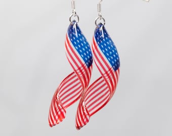 May include: A pair of silver dangle earrings with a twisted design featuring an American flag print. The earrings are red, white, and blue.