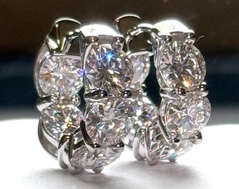 May include: A pair of silver hoop earrings with multiple round, clear gemstones. The earrings are displayed on a white surface, with a dark background. The gemstones sparkle, reflecting light.