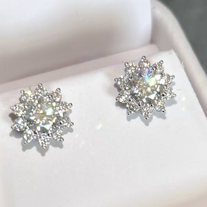 May include: A pair of sparkling diamond stud earrings. Each earring features a large, round, clear center stone surrounded by smaller, clear stones, creating a flower-like design. The earrings are set in a silver-colored metal and are displayed on a white surface.