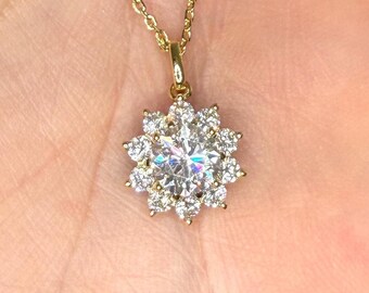May include: A gold necklace featuring a pendant with a large central round-cut clear stone surrounded by a halo of twelve smaller round-cut clear stones. The pendant is attached to a delicate gold chain with a polished, reflective metal finish.