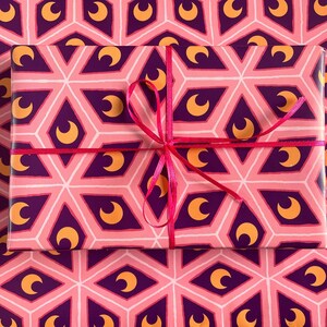 May include: A wrapped gift box with a pink, purple, and gold geometric pattern featuring crescent moons. The box is tied with a bright pink ribbon, and sits on matching wrapping paper.