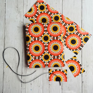 May include: A set of colorful gift wrapping paper with a geometric pattern of black, orange, and white circles. The paper is cut into different sizes and shapes, including a large sheet, a smaller sheet, and a tag.