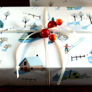 May include: A rectangular gift box wrapped in white paper featuring a winter landscape illustration with houses, trees, and a skier. The package is tied with a thin white ribbon adorned with two red and two orange spherical beads at the center.