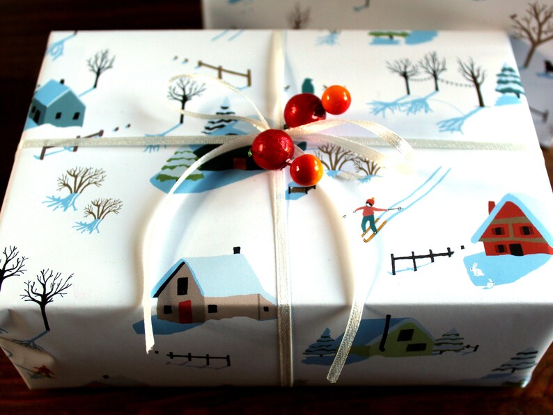 May include: A rectangular gift box wrapped in white paper featuring a winter landscape illustration with houses, trees, and a skier. The package is tied with a thin white ribbon adorned with two red and two orange spherical beads at the center.