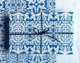 May include: Gift box wrapped in blue and white patterned paper, tied with a black ribbon. The paper features a repeating design of stylized birds and floral elements. The gift box is placed on a matching sheet of wrapping paper.
