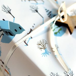 May include: A white gift box wrapped in paper featuring a winter scene with light blue houses, bare trees, and snowmen. The package is tied with a thin white ribbon and decorated with a small, flat, five-pointed wooden star ornament.