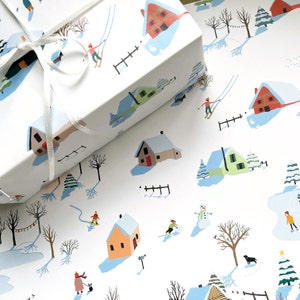 May include: A white gift box wrapped in paper featuring a winter scene with colorful houses, snow-covered trees, people skiing, ice skating, and building a snowman. A thin white ribbon is tied around the box, which sits on matching wrapping paper.