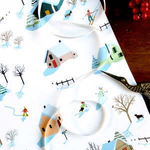 May include: White wrapping paper featuring a repeating winter scene with small houses, bare trees, snowmen, and figures skiing or snowboarding. A thin white ribbon curls across the surface, and gold-colored stork-shaped scissors rest nearby.