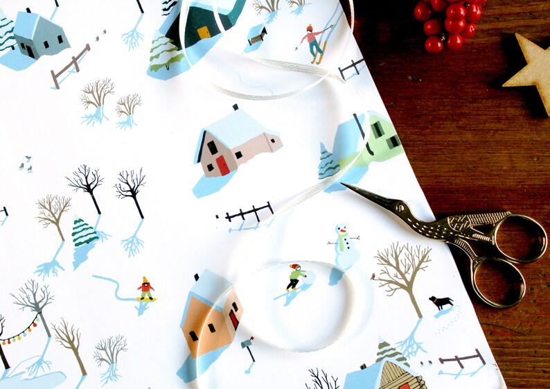 May include: White wrapping paper featuring a repeating winter scene with small houses, bare trees, snowmen, and figures skiing or snowboarding. A thin white ribbon curls across the surface, and gold-colored stork-shaped scissors rest nearby.