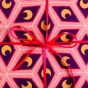 May include: A wrapped gift with pink patterned paper featuring purple diamond shapes with gold crescent moons. A bright red ribbon is tied in a bow around the package, creating a festive and decorative presentation.