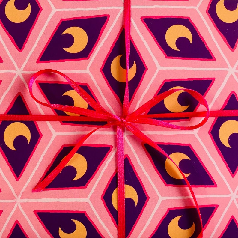 May include: A wrapped gift with pink patterned paper featuring purple diamond shapes with gold crescent moons. A bright red ribbon is tied in a bow around the package, creating a festive and decorative presentation.