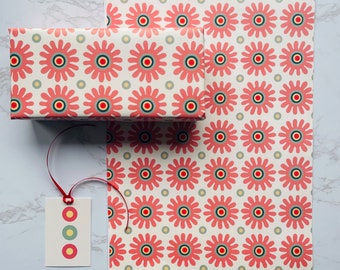 May include: A red and white gift wrap with a repeating pattern of stylized flowers. The gift wrap is folded in half and a red ribbon is tied around it. A gift tag with a yellow, green, and red circle design is attached to the ribbon.