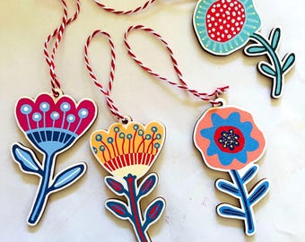 Puede incluir: Cuatro adornos florales de madera plana con cordones para colgar a rayas rojas y blancas. Cada flor presenta combinaciones de colores distintas que incluyen rosa, amarillo, naranja y turquesa, con pétalos, tallos y hojas pintados.
