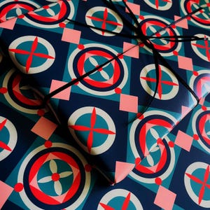May include: Gift-wrapped present with a geometric pattern in teal, red, and white on a navy blue background. The design includes circles, crosses, and squares. A black ribbon is tied around the gift.