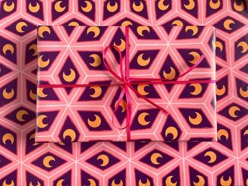 May include: A wrapped gift box with a pink, purple, and gold geometric pattern featuring crescent moons. The box is tied with a bright pink ribbon, and sits on matching wrapping paper.
