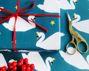 May include: A teal gift box wrapped with a thin red ribbon and bow, featuring a repeating pattern of white swans wearing small yellow crowns and scattered yellow stars. Beside the box are gold-colored metal stork scissors and red berries.