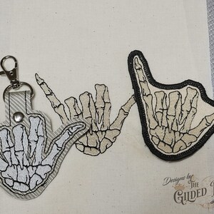 Può includere: Due toppe ricamate con mani scheletriche che fanno il gesto 'rock on'. Le toppe sono beige con contorni neri e sono su uno sfondo di tessuto beige.