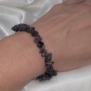 May include: A dark blue chip stone bracelet is worn on a wrist. The bracelet is made of small, irregularly shaped stones. The background is a white, silky fabric.