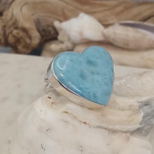 Könnte beinhalten: Ein Silberring mit einem herzförmigen, hellblauen Edelstein. Der Edelstein hat ein marmoriertes Aussehen und ist in einem Silberband gefasst. Der Ring wird auf einer weißen Oberfläche präsentiert, um das Design des Schmucks hervorzuheben.