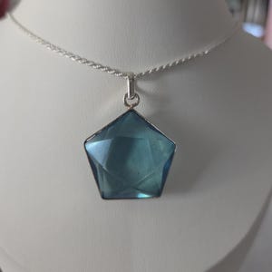 May include: A pentagonal blue gemstone pendant set in a silver-toned metal frame, suspended from a thin, twisted silver-toned chain. The translucent blue stone features geometric facets that catch the light, creating a clear, sharp shape.