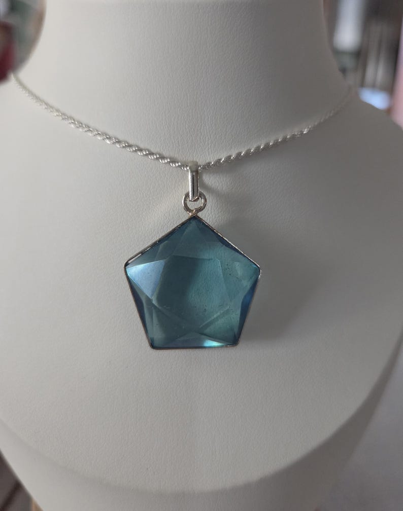 May include: A pentagonal blue gemstone pendant set in a silver-toned metal frame, suspended from a thin, twisted silver-toned chain. The translucent blue stone features geometric facets that catch the light, creating a clear, sharp shape.