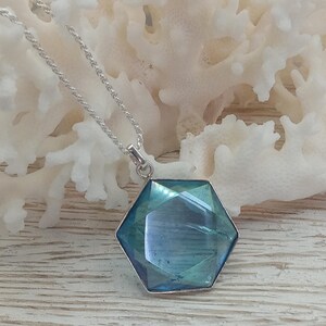 May include: A hexagonal pendant featuring a faceted blue-green gemstone set in a silver-toned metal frame, suspended from a delicate silver-toned rope chain. The pendant displays a reflective surface with deep teal and azure tones.