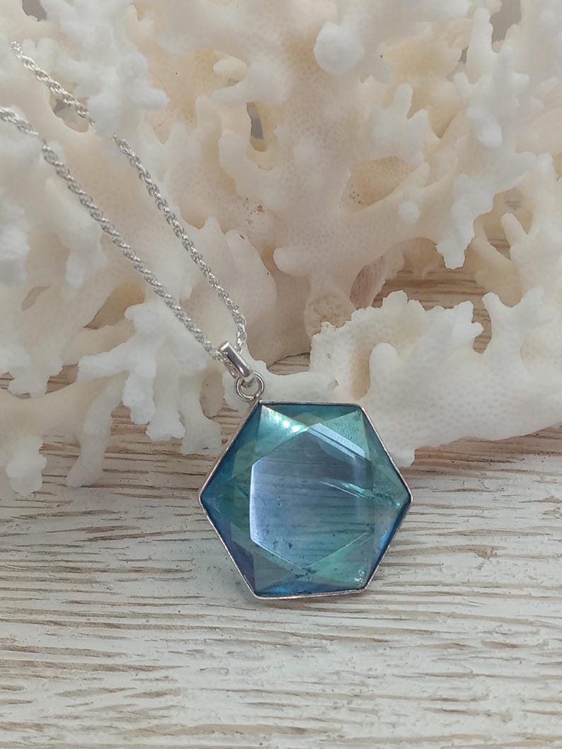 May include: A hexagonal pendant featuring a faceted blue-green gemstone set in a silver-toned metal frame, suspended from a delicate silver-toned rope chain. The pendant displays a reflective surface with deep teal and azure tones.