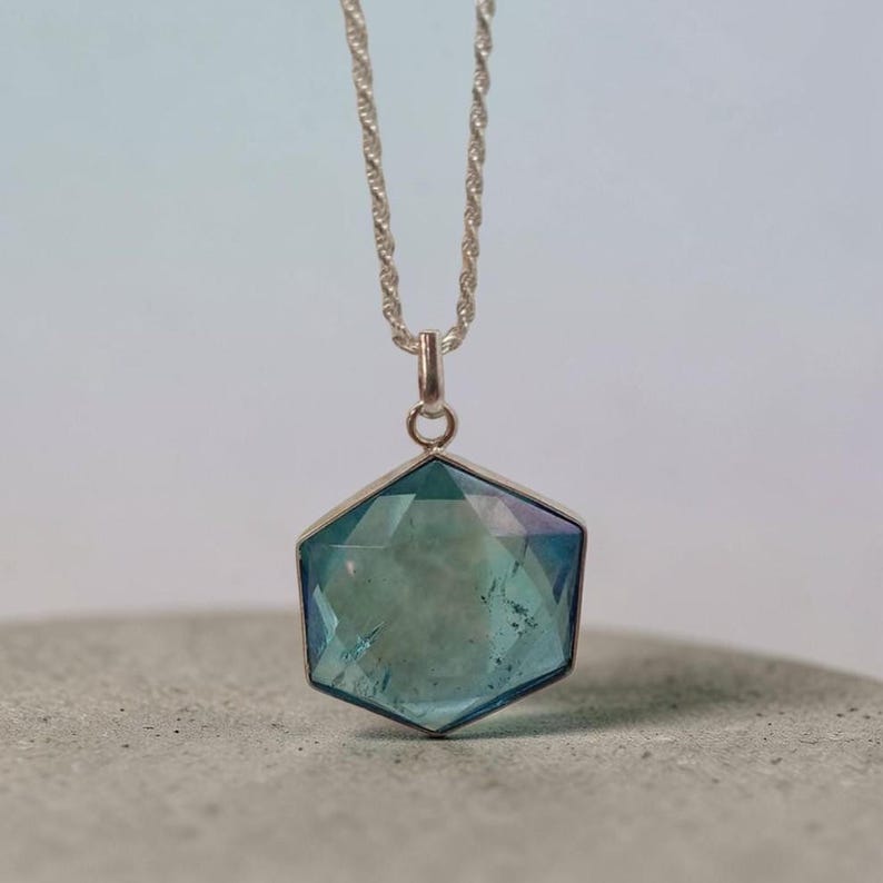 May include: A silver necklace featuring a hexagonal pendant with a faceted, translucent blue-green gemstone. The pendant is suspended from a thin, twisted silver chain, showcasing a clear, polished surface with visible internal inclusions.