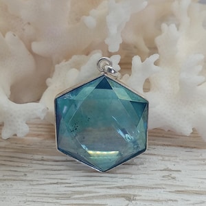 May include: A hexagonal pendant featuring a faceted, translucent blue-green stone set in a silver-toned metal frame. The pendant includes a small circular bail at the top for attachment, showcasing a geometric cut with reflective surfaces.