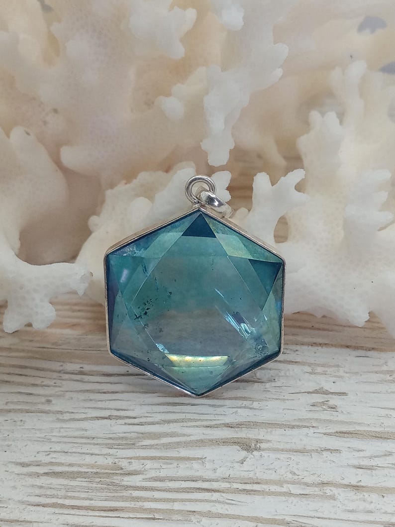 May include: A hexagonal pendant featuring a faceted, translucent blue-green stone set in a silver-toned metal frame. The pendant includes a small circular bail at the top for attachment, showcasing a geometric cut with reflective surfaces.