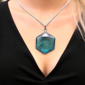 May include: A silver-toned rope chain necklace featuring a large, faceted, hexagonal teal-colored pendant. The pendant has a smooth, polished surface with subtle internal inclusions and is suspended from a simple silver-toned metal bail.