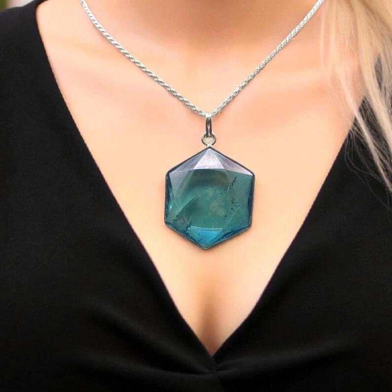 May include: A silver-toned rope chain necklace featuring a large, faceted, hexagonal teal-colored pendant. The pendant has a smooth, polished surface with subtle internal inclusions and is suspended from a simple silver-toned metal bail.