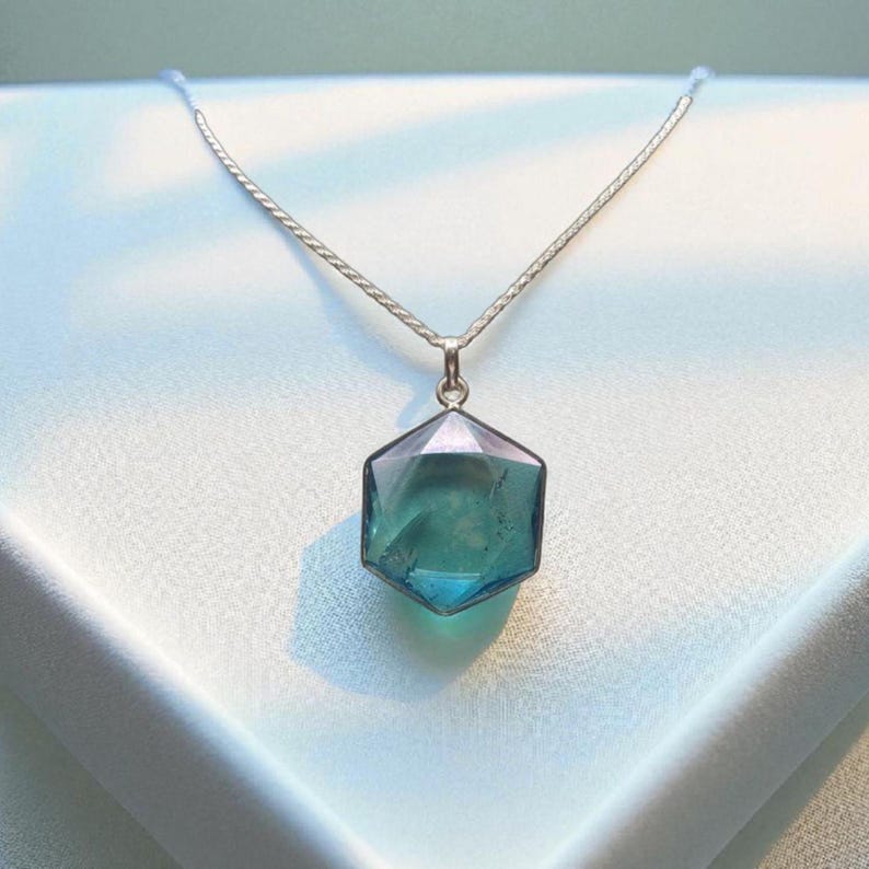 May include: A silver-toned chain necklace featuring a hexagonal pendant with a translucent, multifaceted teal and blue gemstone. The pendant is suspended from a delicate metal chain, showcasing a clear, polished surface with internal depth.