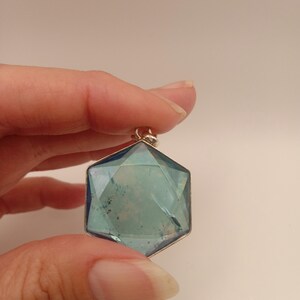 May include: A hexagonal pendant featuring a translucent blue faceted gemstone set in a thin silver-toned metal frame. The stone displays internal inclusions and a geometric cut with a small metal bail attached to the top point for hanging.