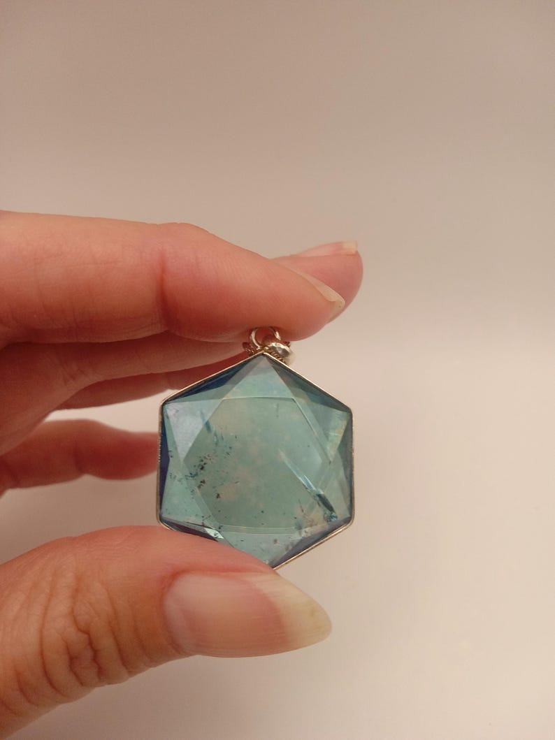 May include: A hexagonal pendant featuring a translucent blue faceted gemstone set in a thin silver-toned metal frame. The stone displays internal inclusions and a geometric cut with a small metal bail attached to the top point for hanging.