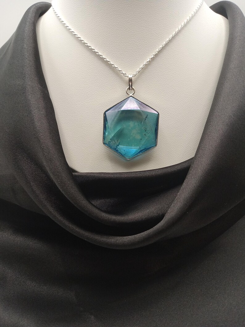 May include: A hexagonal pendant featuring a translucent blue and teal gemstone with a faceted surface, suspended from a thin silver-toned chain. The pendant displays internal inclusions and a polished finish, reflecting light across its geometric edges.