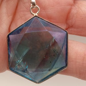 May include: A hexagonal pendant featuring a faceted, translucent blue and purple stone set in a silver-toned metal frame. The pendant hangs from a silver-toned rope chain, displaying a geometric cut with visible internal inclusions and light reflections.