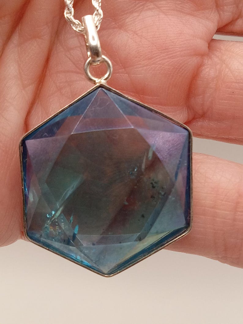 May include: A hexagonal pendant featuring a faceted, translucent blue and purple stone set in a silver-toned metal frame. The pendant hangs from a silver-toned rope chain, displaying a geometric cut with visible internal inclusions and light reflections.