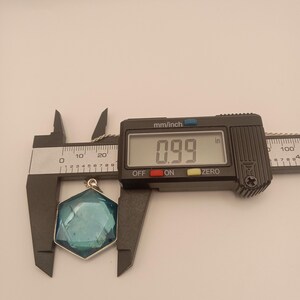 May include: A hexagonal blue pendant with a faceted surface held by a black digital caliper. The display shows a measurement of 0.99 inches. The pendant is attached to a thin silver-colored chain visible at the top of the frame.