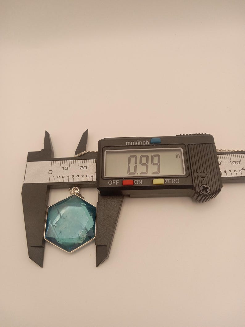 May include: A hexagonal blue pendant with a faceted surface held by a black digital caliper. The display shows a measurement of 0.99 inches. The pendant is attached to a thin silver-colored chain visible at the top of the frame.
