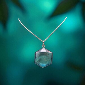 May include: A silver-toned necklace featuring a hexagonal pendant with a translucent, light blue gemstone. The pendant is suspended from a delicate, thin silver-toned chain, showcasing a clean, geometric design with smooth, polished edges.