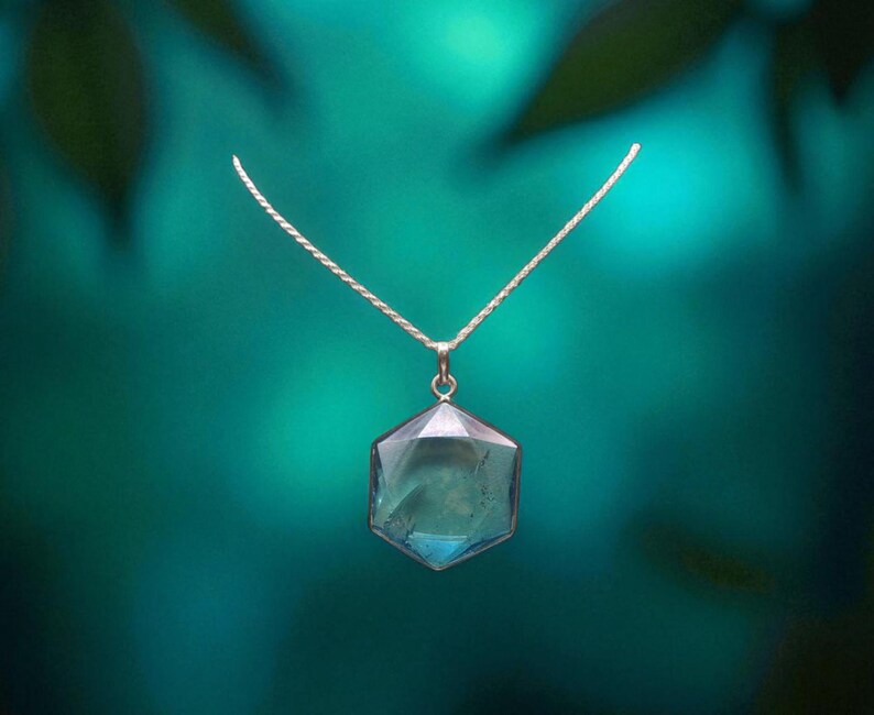 May include: A silver-toned necklace featuring a hexagonal pendant with a translucent, light blue gemstone. The pendant is suspended from a delicate, thin silver-toned chain, showcasing a clean, geometric design with smooth, polished edges.