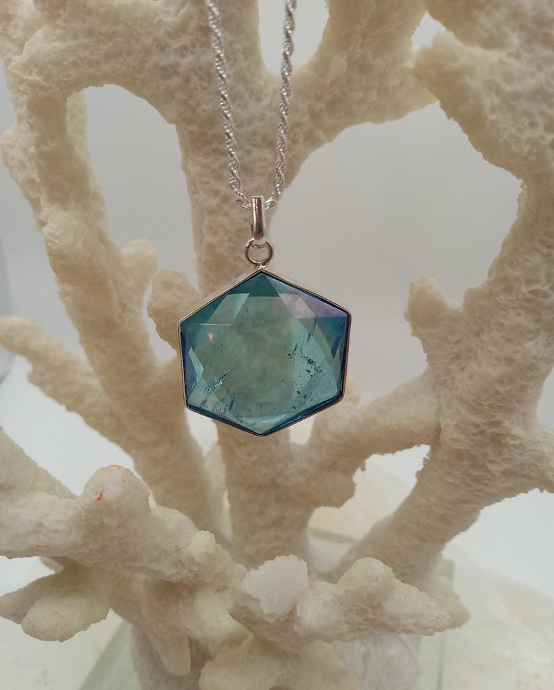 May include: A silver necklace with a hexagonal pendant. The pendant has a faceted, teal-colored gemstone with a silver border. The necklace is displayed against a white, coral-like background.