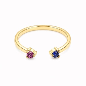 May include: A gold-colored open ring with a magenta gemstone on one end and a blue gemstone on the other. The ring is set against a white background, highlighting the ring's design and the colors of the gemstones.