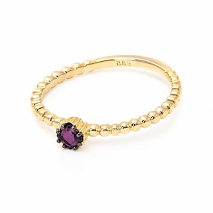 Op de afbeelding: Een gouden ring met een getextureerde band en een kleine, ronde, paarse edelsteen. De ring heeft een kralenontwerp langs de band en een donkere zetting voor de edelsteen. De achtergrond is wit.