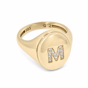 May include: A gold signet ring featuring a polished oval face with a letter M inlaid with small clear stones. The interior of the band is clearly stamped with the number 585, indicating the gold purity of the metal used for this jewelry piece.