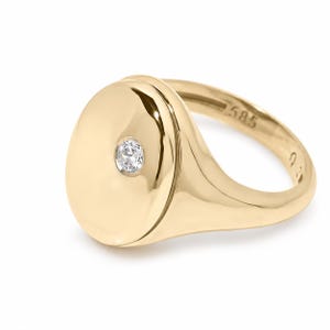 May include: A polished gold signet ring featuring a smooth, oval-shaped face with a single, small, round, clear gemstone set off-center. The interior of the band is clearly engraved with the hallmark 585, indicating the gold purity of the ring.