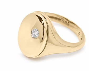 May include: A polished gold signet ring featuring a smooth, oval-shaped face with a single, small, round, clear gemstone set off-center. The interior of the band is clearly engraved with the hallmark 585, indicating the gold purity of the ring.